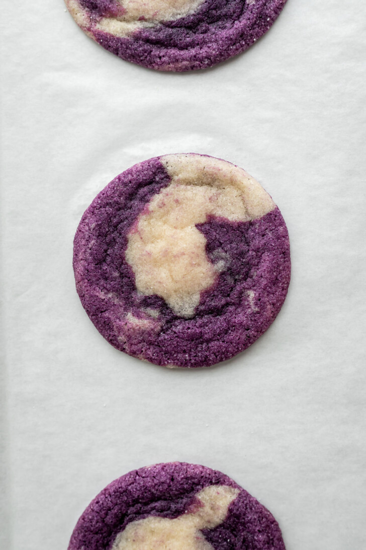 A close-up of three round ube marble cookies with a marbled pattern of purple and white, displayed on a sheet of white parchment paper.