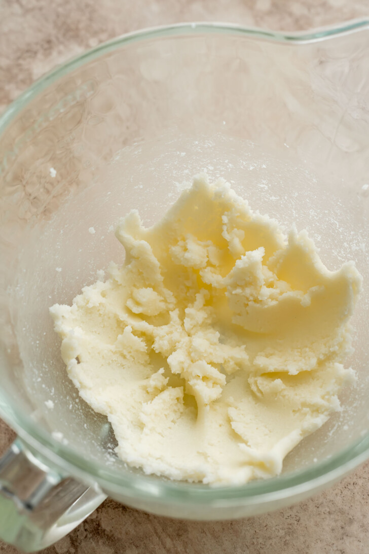A glass mixing bowl containing creamed butter and sugar on a light brown countertop, with the mixture smooth and slightly fluffy in texture.