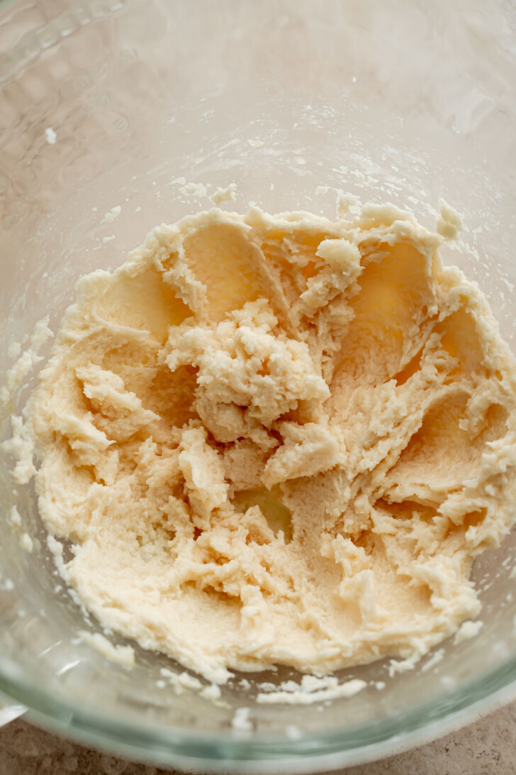 A close-up of a glass mixing bowl containing creamed butter and sugar, showing a light, fluffy, pale mixture with a whipped texture.