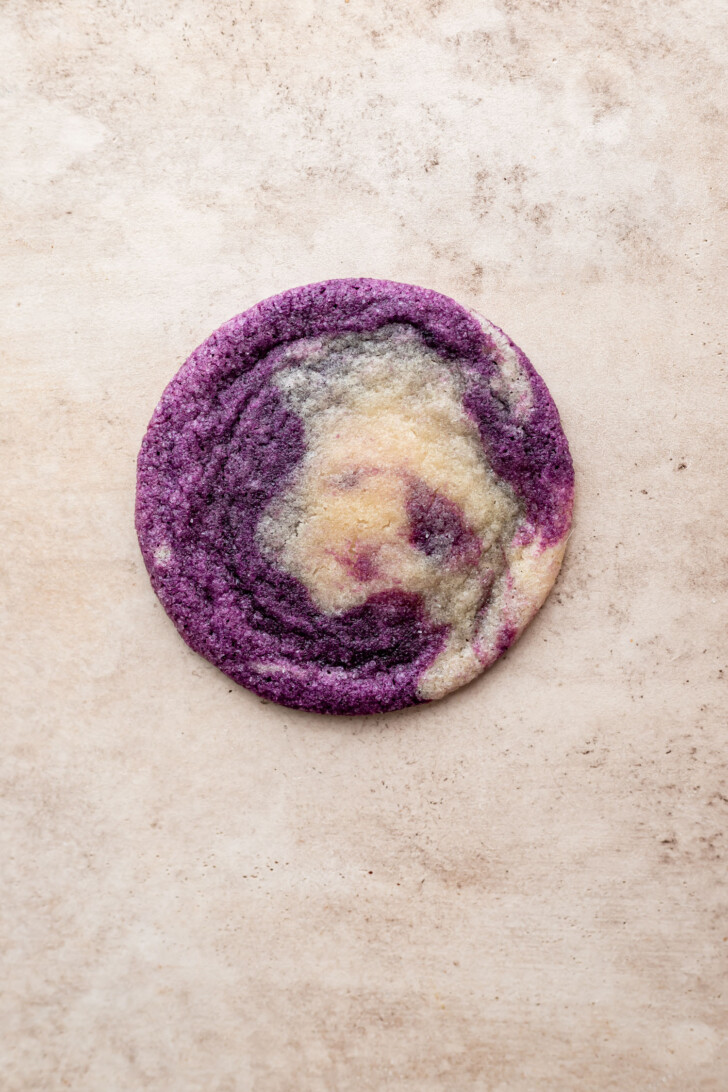 A round cookie with a marbled pattern of vibrant purple and creamy white swirls, placed on a light beige textured surface.