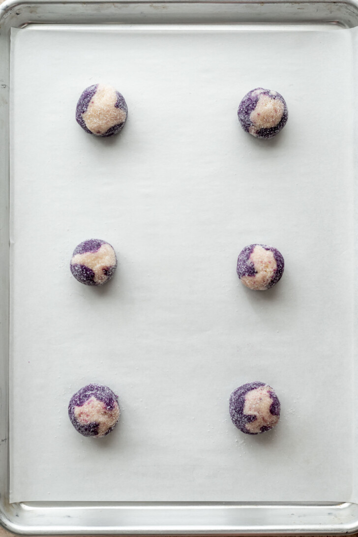 Six round, marbled purple and white cookie dough balls are evenly spaced on a parchment-lined baking sheet, ready to be baked.