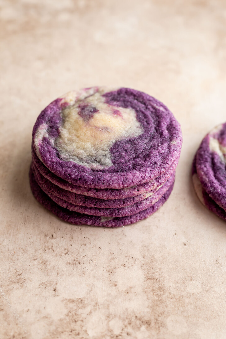 A stack of vibrant purple and white ube marble cookies sits on a light beige surface, with one additional cookie partially visible to the right.