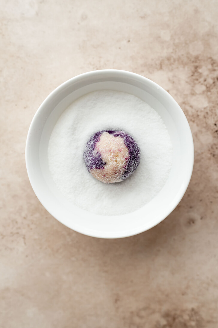 A round, sugar-coated purple dough ball sits in a white bowl filled with granulated sugar, placed on a light beige surface.