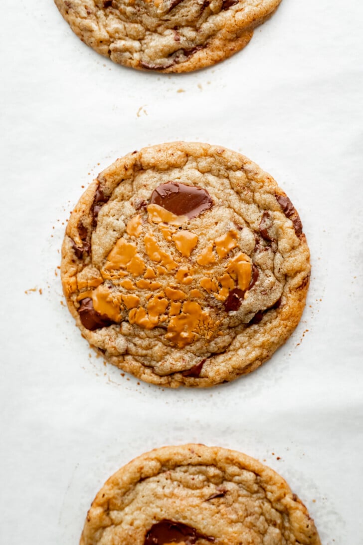 A close-up of a chewy chocolate chip cookie with melted caramel and a swirl of biscoff cookie butter on top, resting on white parchment paper. Partial cookies are visible at the top and bottom edges of the image.