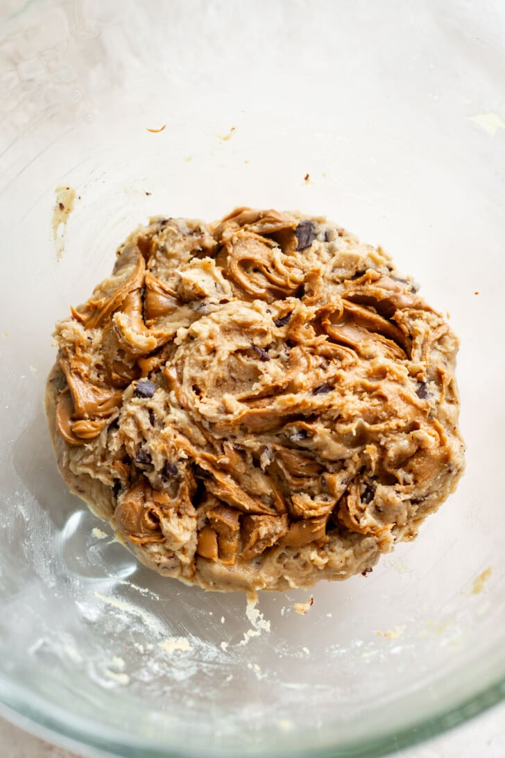 A close-up of cookie dough with visible swirls of peanut butter, biscoff cookie butter, and chocolate chips in a clear glass mixing bowl.
