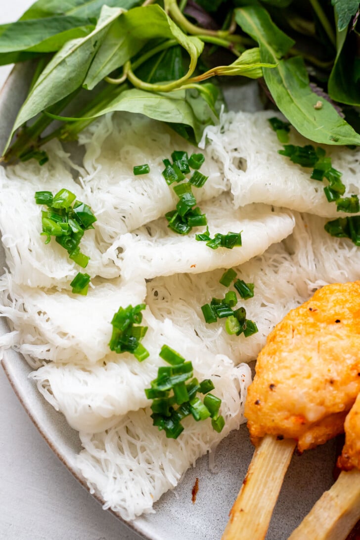 A close-up of Vietnamese banh hoi rice vermicelli noodles topped with chopped green onions, served with fresh herbs and orange grilled chao tom shrimp skewers on a plate.
