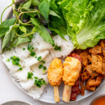 A plate of banh hoi with fresh lettuce, mixed herbs, rice vermicelli noodles topped with scallions, grilled meat, and shrimp paste on sugarcane, served with a flavorful Vietnamese dipping sauce on the side.