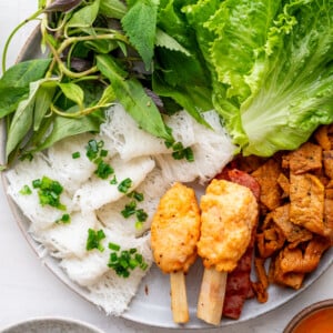 A plate of banh hoi with fresh lettuce, mixed herbs, rice vermicelli noodles topped with scallions, grilled meat, and shrimp paste on sugarcane, served with a flavorful Vietnamese dipping sauce on the side.
