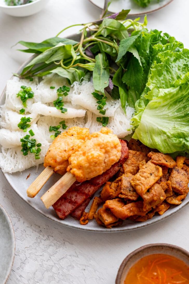 A plate with Vietnamese ingredients: fresh lettuce, herbs, banh hoi rice vermicelli, grilled nem nuong pork, chao tom skewered shrimp paste, and slices of Vietnamese sausage, served with a small bowl of orange dipping sauce on the side.