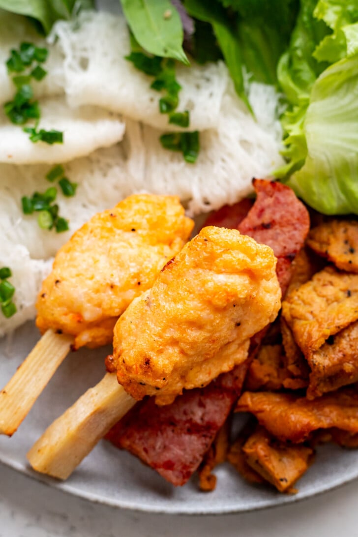 Close-up of a plate with chao tom (grilled shrimp paste on sugarcane), nem nuong, fresh greens, and banh hoi vermicelli noodles, beautifully garnished with scallion oil.