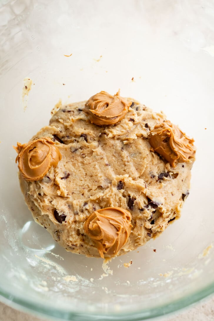 A glass mixing bowl containing cookie dough with chocolate chunks and four dollops of creamy biscoff cookie butter placed on top of the dough.