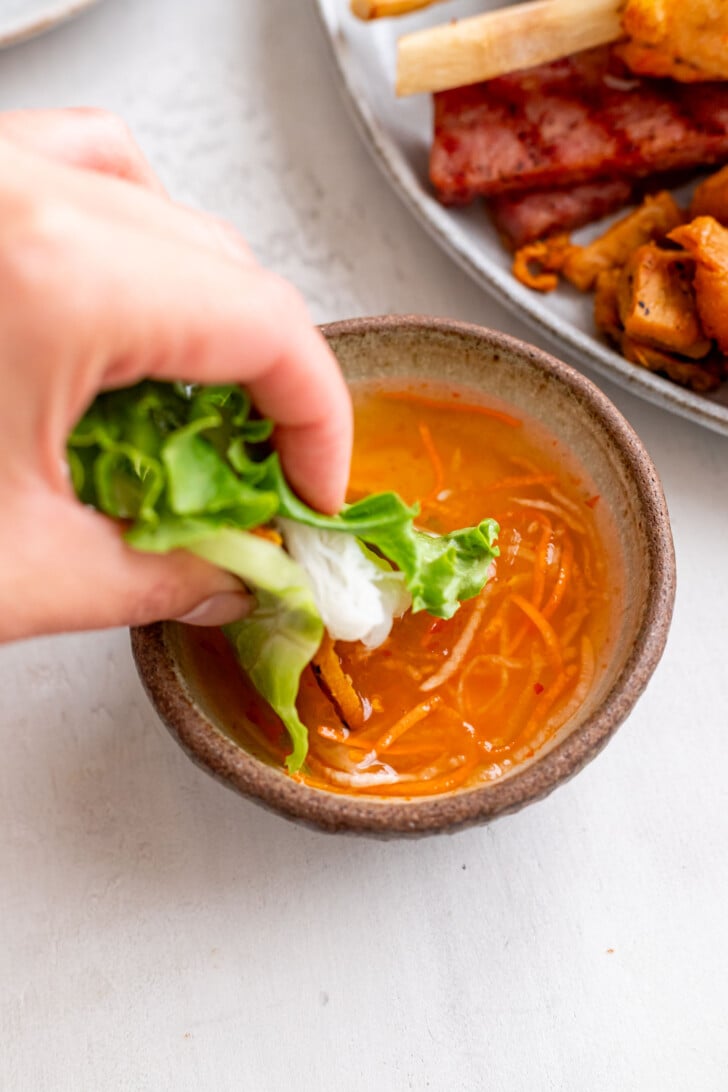 A hand dips a lettuce wrap filled with lemongrass pork into a small bowl of orange nuoc cham sauce with shredded vegetables, with a plate of grilled meat, banh hoi, and vegetables in the background.