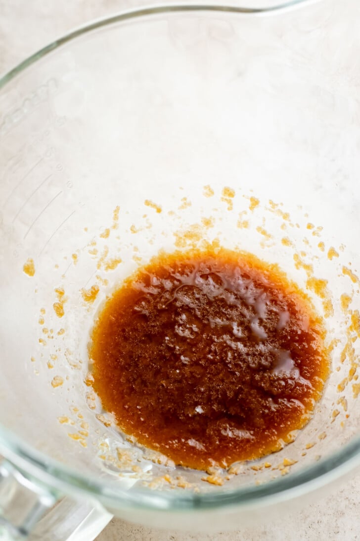 A close-up of a glass mixing bowl containing a mixture of brown sugar and melted butter on a light-colored surface.