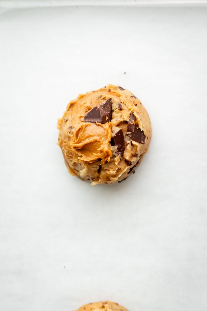A ball of unbaked cookie dough with chocolate chunks and a dollop of biscoff cookie butter on top, resting on a sheet of parchment paper.
