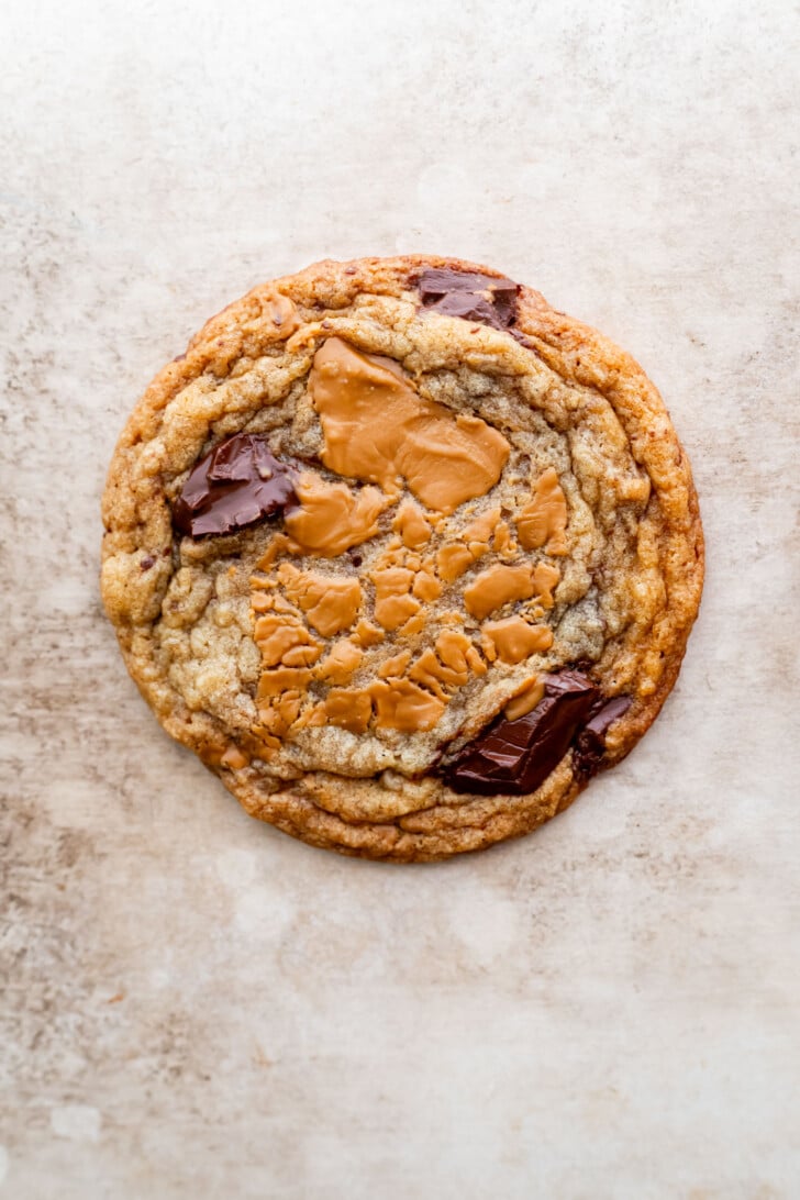 A large, round cookie with a golden-brown edge, chunks of dark chocolate, and a melted swirl of biscoff cookie butter in the center, set on a light, textured surface.