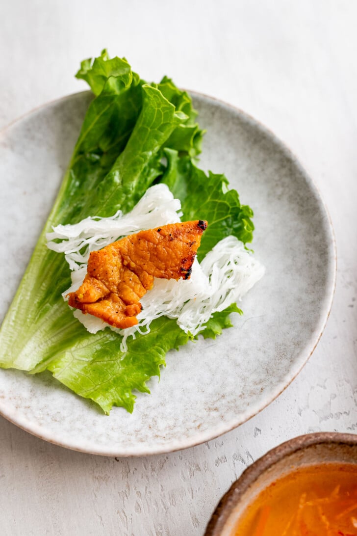 A plate with a fresh green lettuce leaf topped with white rice vermicelli noodles, tender lemongrass pork, and a bowl of orange dipping sauce partially visible at the edge, highlights this flavorful banh hoi dish.