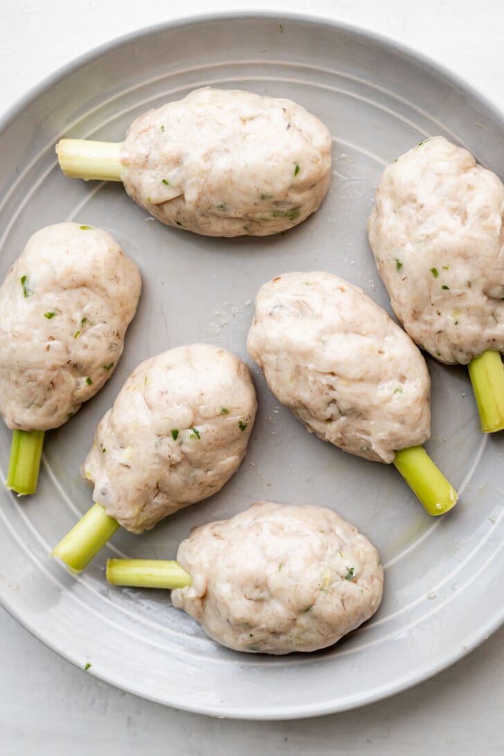 Six uncooked shrimp and pork paste skewers formed around lemongrass sticks and arranged on a round gray plate.