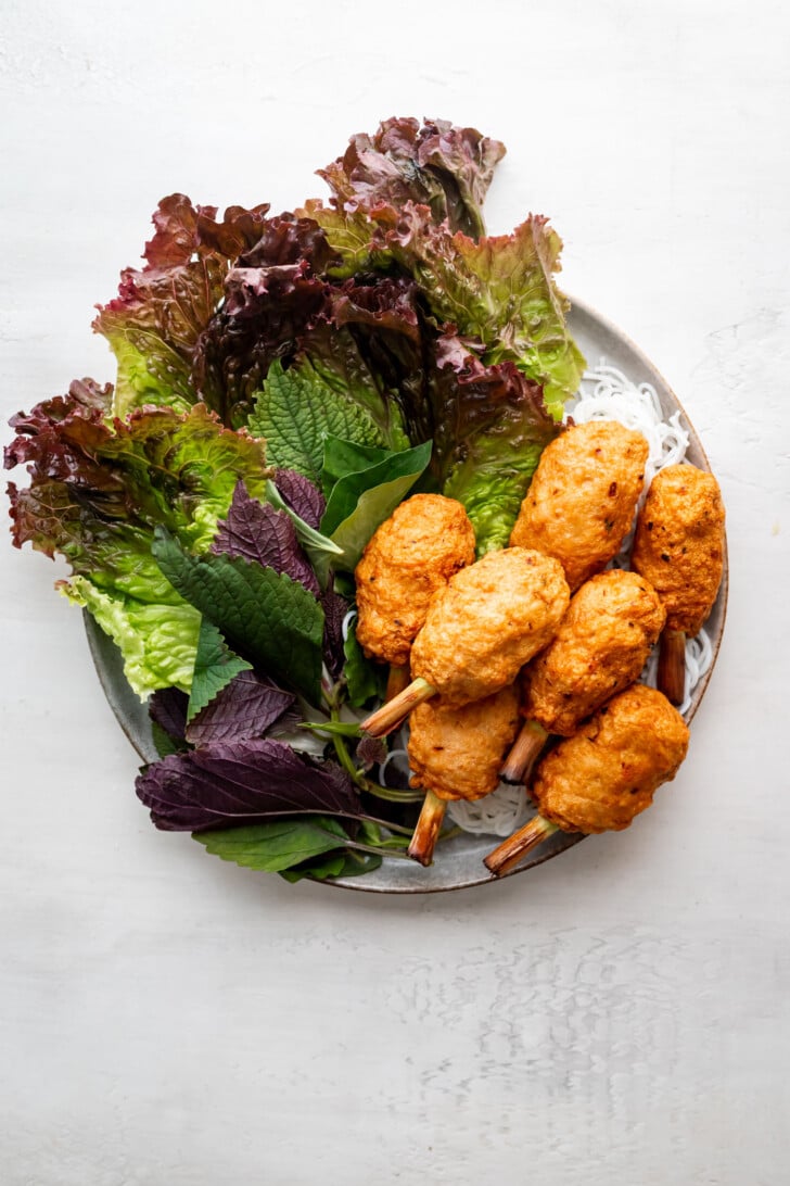 A plate of chao tom—shrimp paste on lemongrass sticks—served with fresh green and purple leafy herbs, lettuce, and a small portion of white vermicelli noodles.