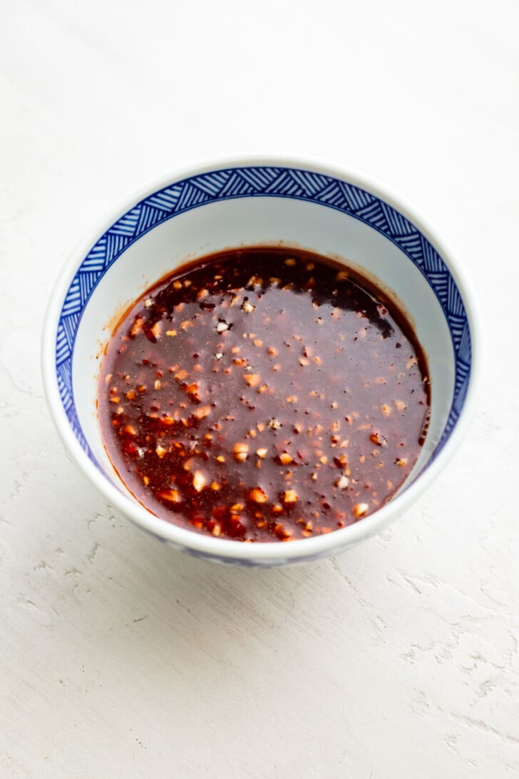 A blue and white patterned bowl filled with a reddish-brown, chunky char siu sauce sits on a light textured surface. The sauce contains visible minced garlic.