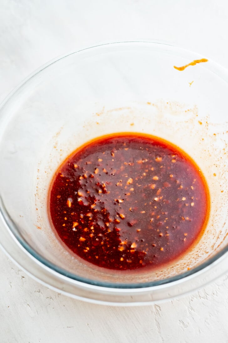 A clear glass bowl filled with char siu sauce&mdash;a reddish-brown, slightly chunky marinade&mdash;sits on a white surface. The sauce appears to contain spices and small bits of ingredients.