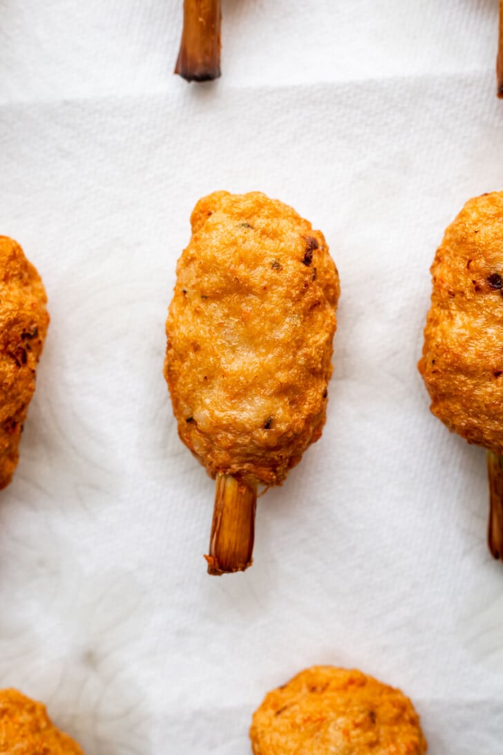 Golden-brown, crispy fried lollipop-shaped chao tom snacks with exposed lemongrass sticks, arranged on a white paper towel to absorb excess oil.