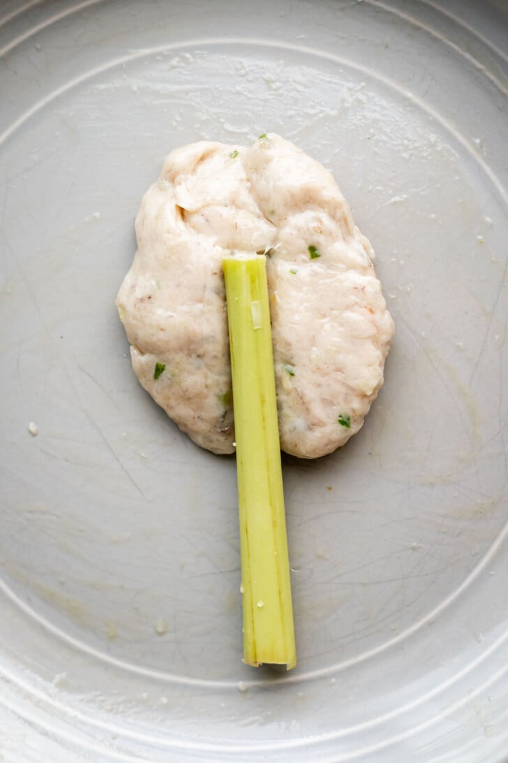 A mound of seasoned shrimp and pork paste shaped on a circular gray plate with a lemongrass stick placed lengthwise in the center.