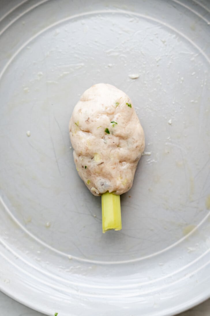 A piece of uncooked, herb-speckled shrimp and pork paste is molded around the top of a lemongrass stick, placed on a round, gray plate.