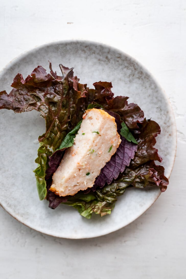 A piece of chao tom garnished with chopped herbs sits on top of leafy red and green lettuce, served on a round, gray ceramic plate against a white background.