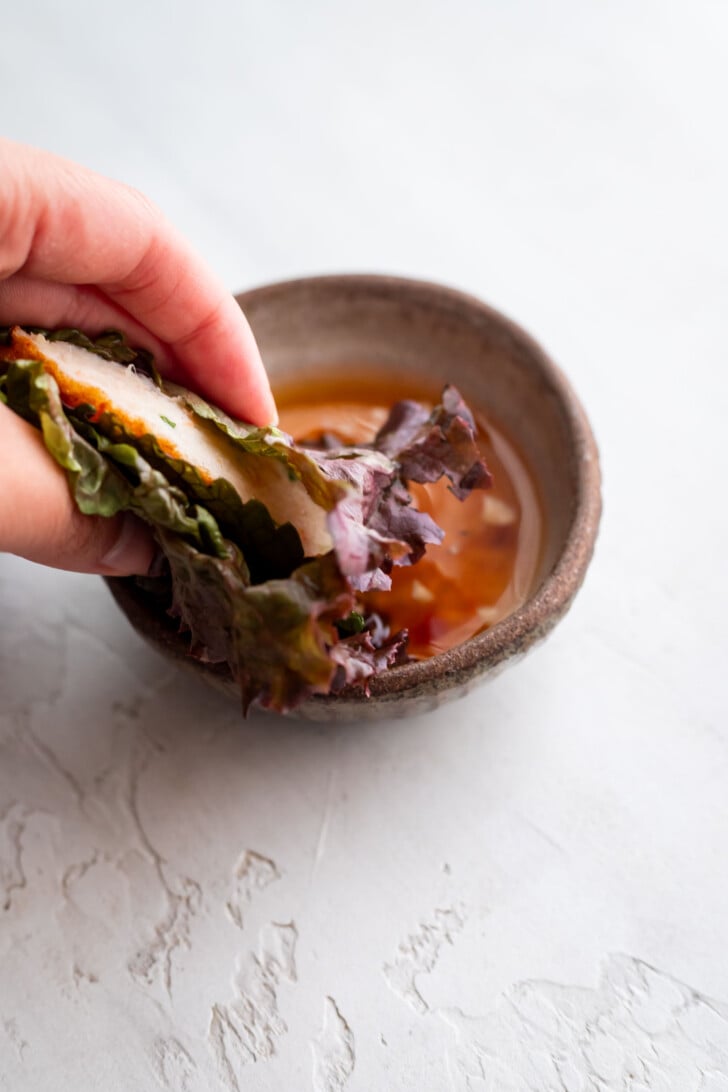 A hand holds a lettuce wrap filled with chao tom and vegetables, dipping it into a small bowl of nuoc cham on a light textured surface.