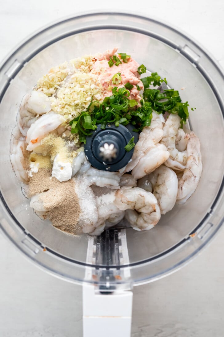 Raw shrimp, chopped green onions, minced garlic, ground spices, and pork paste are placed together in a food processor bowl, viewed from above, ready to be blended.