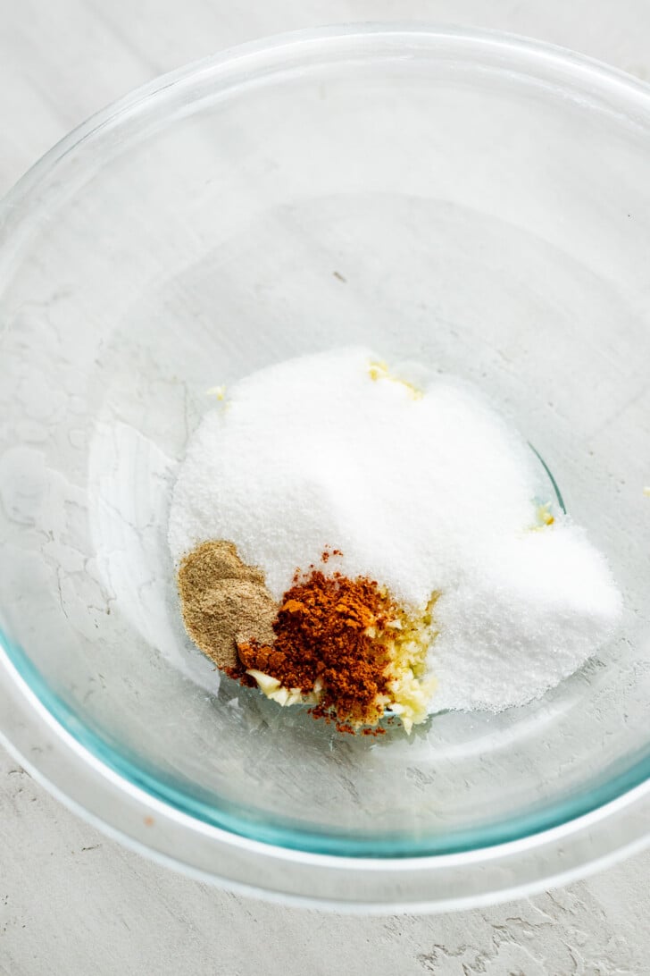 A clear glass bowl containing separate piles of white sugar, ground black pepper, minced garlic, and five spice powder on a light-colored surface.