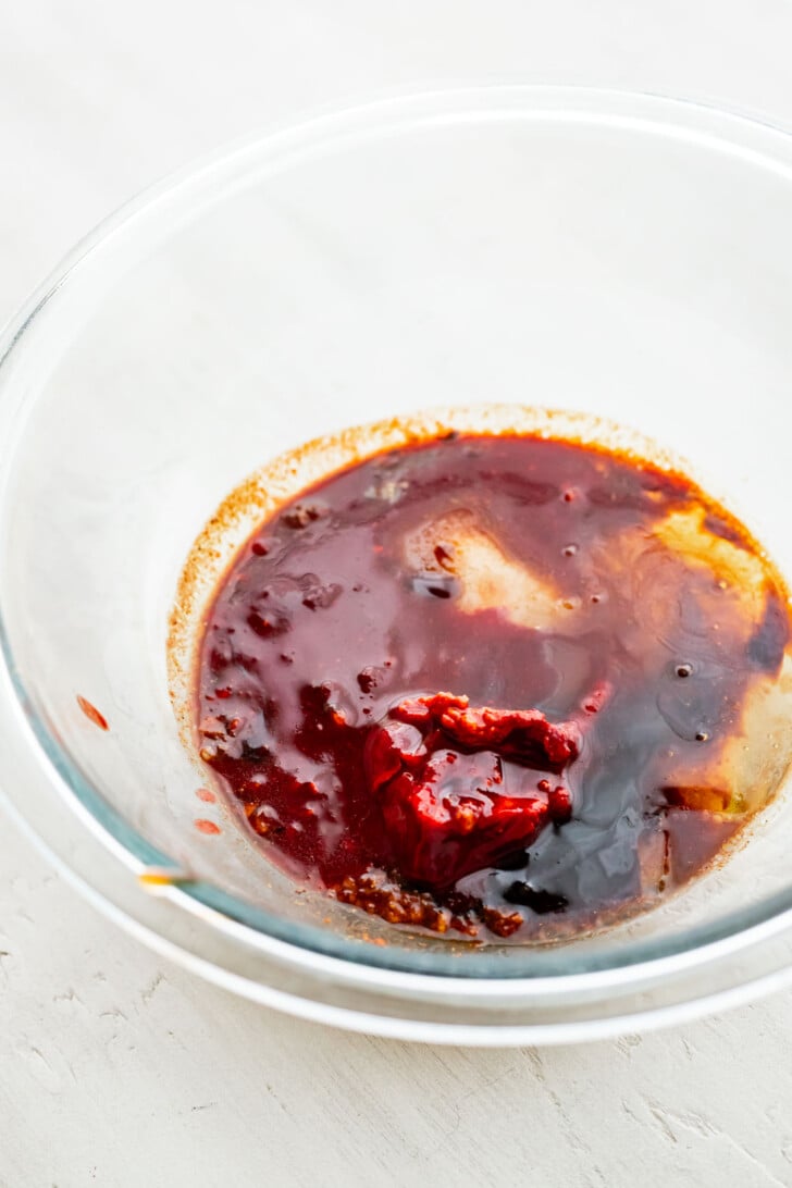 A clear glass bowl containing a mix of red, brown, and yellow sauces or pastes, possibly including chili paste, soy sauce, and oil, on a light-colored surface.