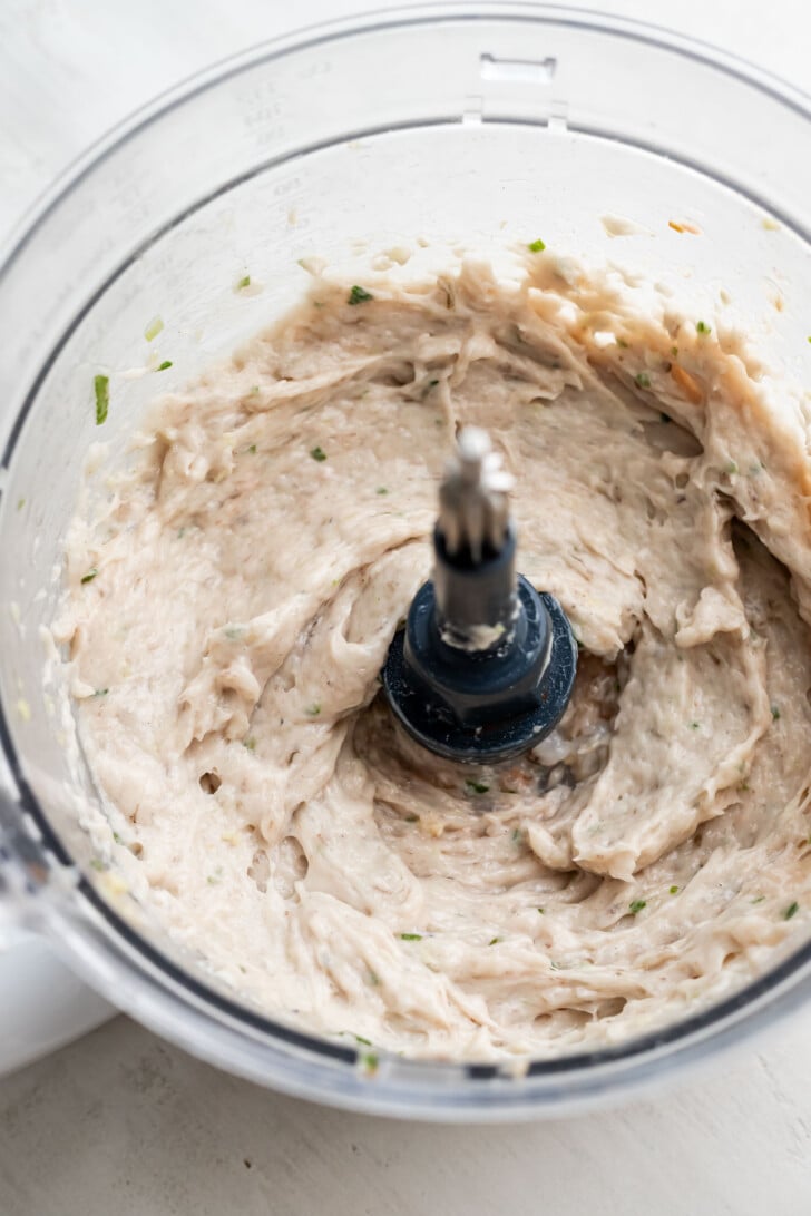 A creamy, blended mixture with visible green herb flecks inside a food processor bowl, viewed from above. The mixture has a thick, smooth texture and the processor blade is centered in the image.