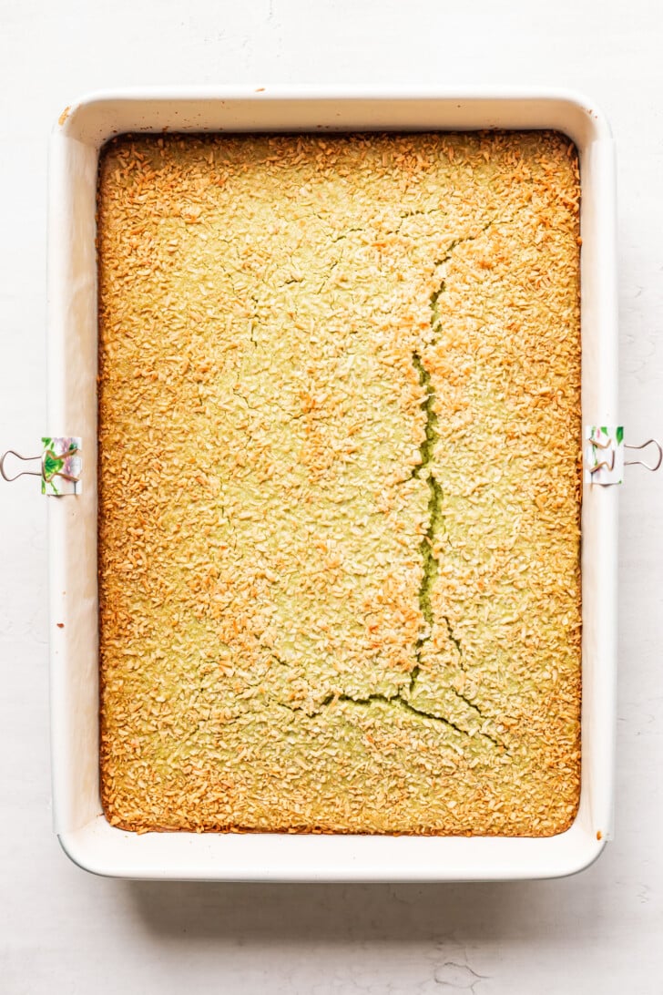 A rectangular baking dish filled with a golden-brown baked cake, topped with toasted coconut flakes and showing a large crack running through the center.