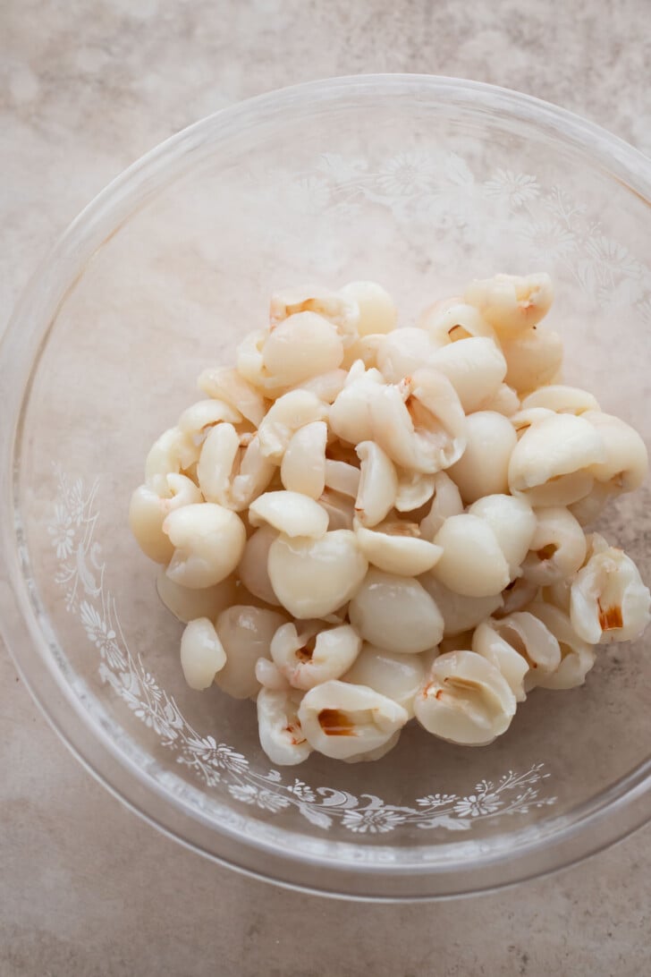 A clear glass bowl filled with peeled and pitted lychee fruit sits on a light-colored countertop. The translucent white fruit pieces are clustered together inside the bowl.