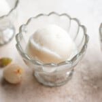A scoop of white lychee sorbet in a scalloped glass bowl sits on a light surface, with a few small fruits nearby