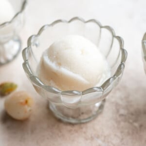 A scoop of white lychee sorbet in a scalloped glass bowl sits on a light surface, with a few small fruits nearby
