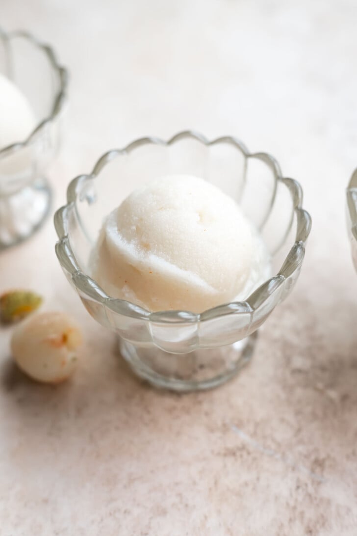 A scoop of white lychee sorbet in a scalloped glass bowl sits on a light surface, with a few small fruits nearby