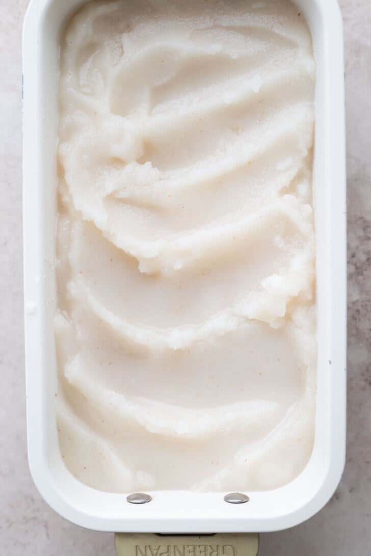 A rectangular white container filled with smooth, creamy white lychee sorbet, with swirled ridges on the surface.