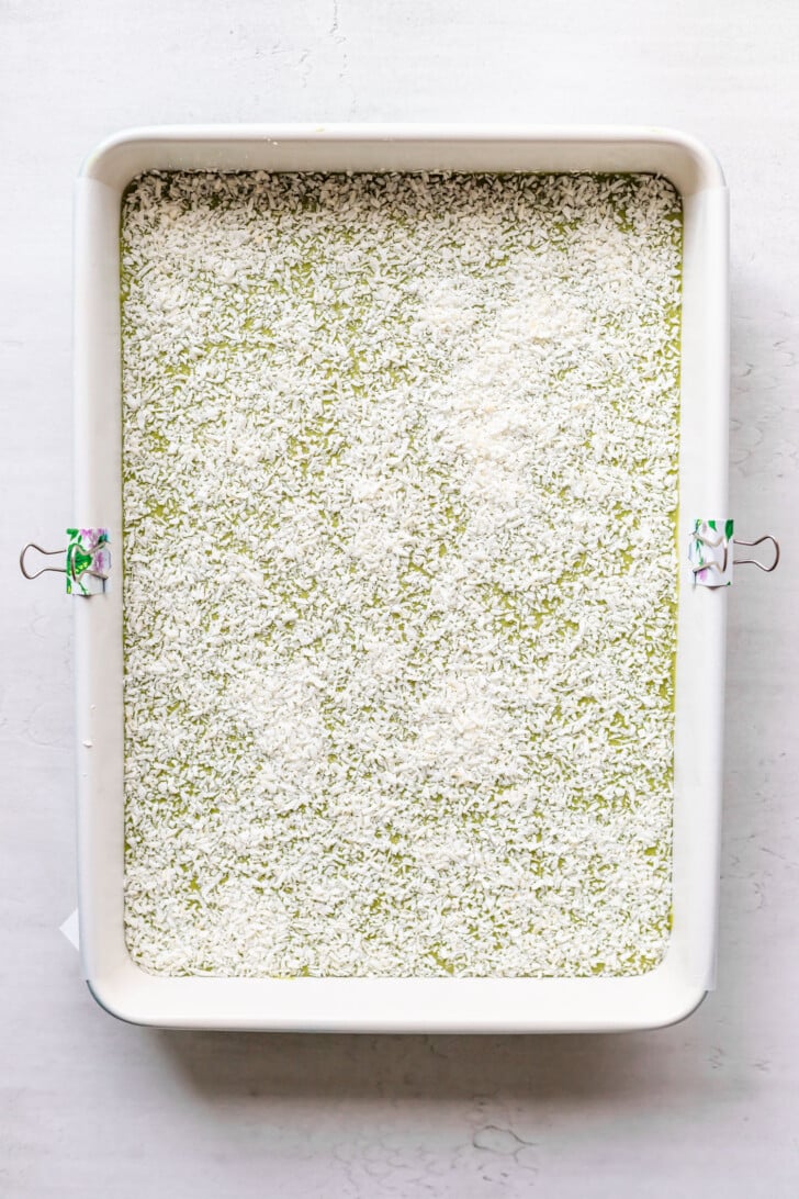 A rectangular baking dish filled with a green mixture topped with a layer of shredded coconut, secured with metal binder clips on the sides, sits on a light surface.