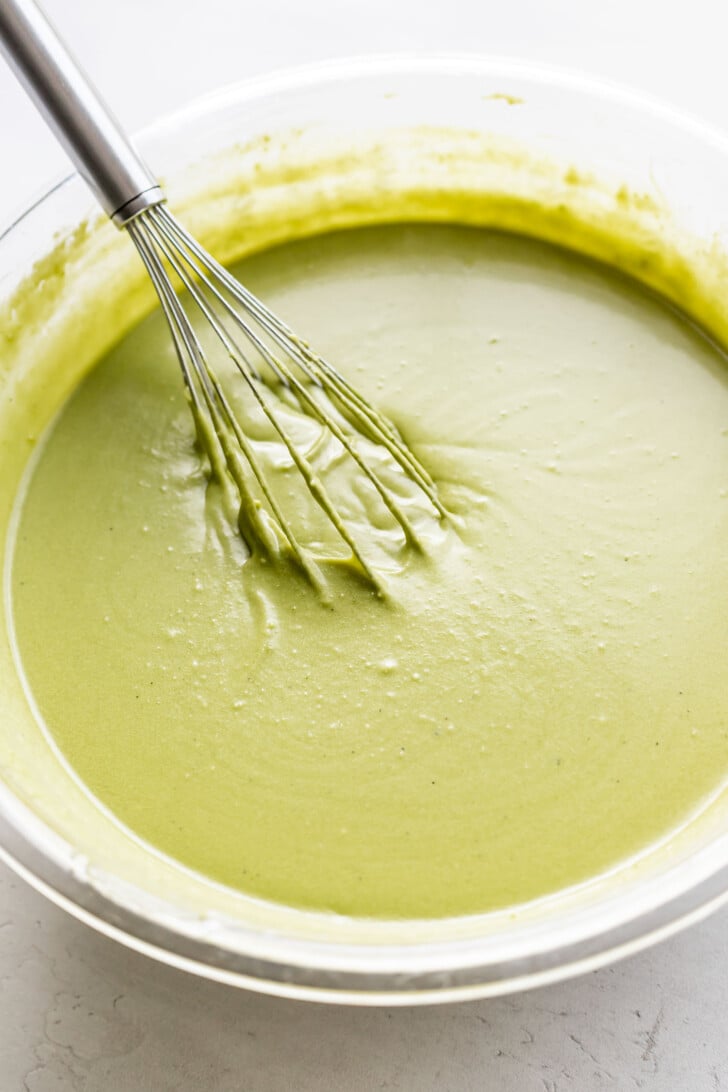 A metal whisk rests in a glass bowl filled with smooth, light green batter or mixture, sitting on a white surface.