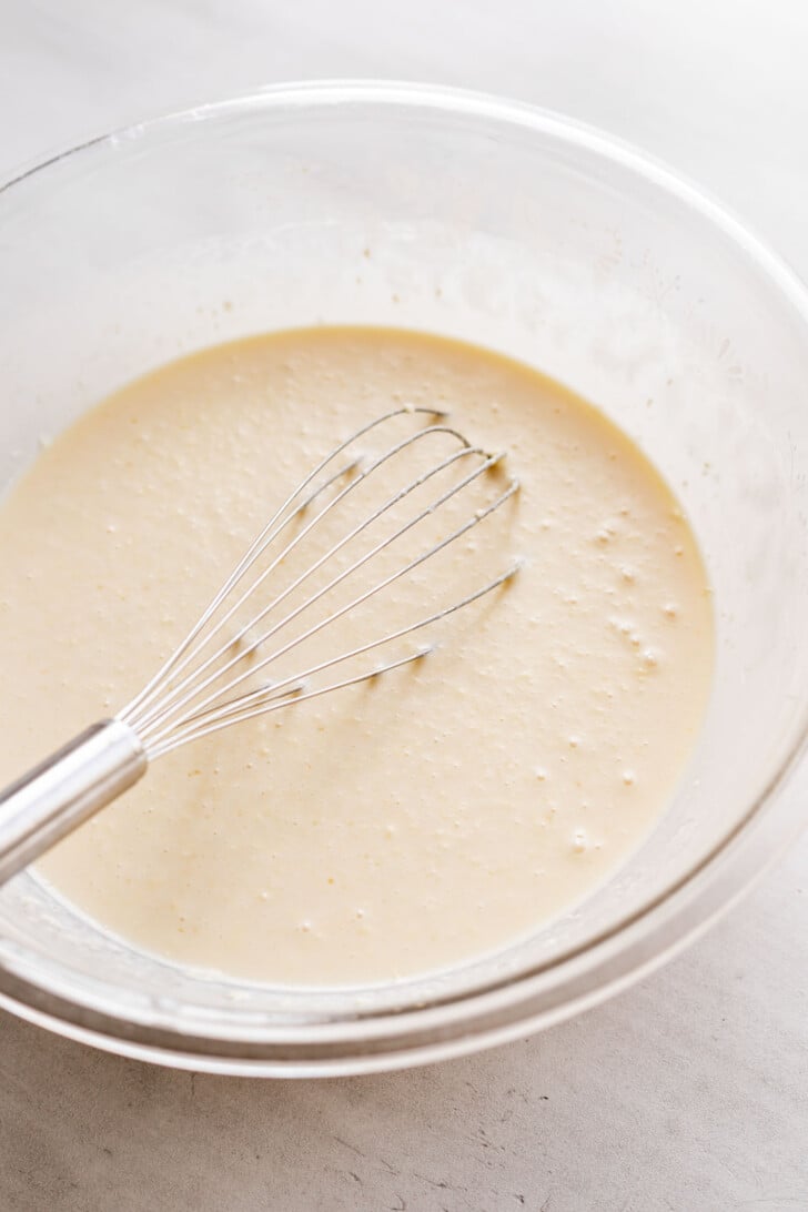 A metal whisk rests in a glass bowl filled with smooth, light-colored batter on a white surface.