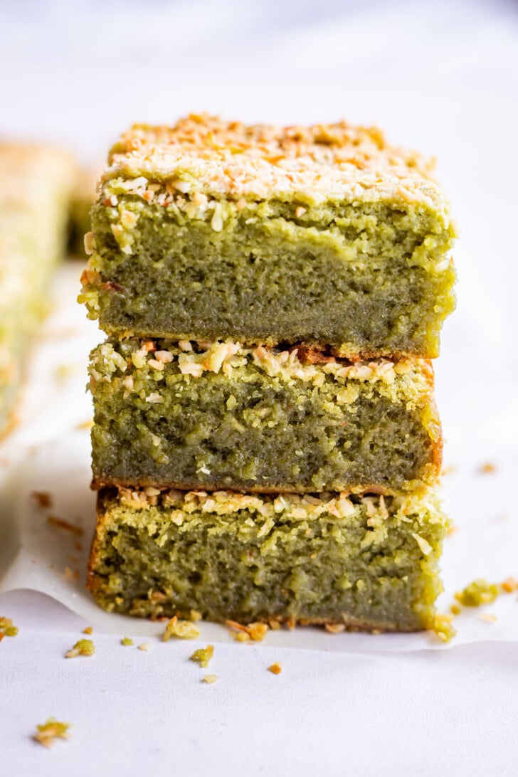 Three pieces of green matcha butter mochi coconut brownies are stacked on top of each other, showing a moist texture with a golden coconut topping, set on a white surface with a few crumbs around.