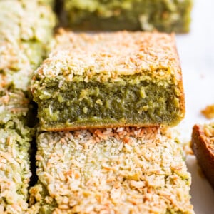 A close-up of green matcha butter mochi bars topped with toasted shredded coconut, with one butter mochi bar on top showing a soft, moist interior.