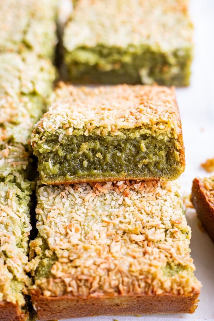 A close-up of green matcha butter mochi bars topped with toasted shredded coconut, with one butter mochi bar on top showing a soft, moist interior.