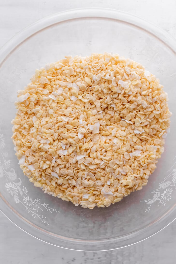 A clear glass bowl filled with crumbled crispy rice cereal and coconut flakes sit on a white surface.