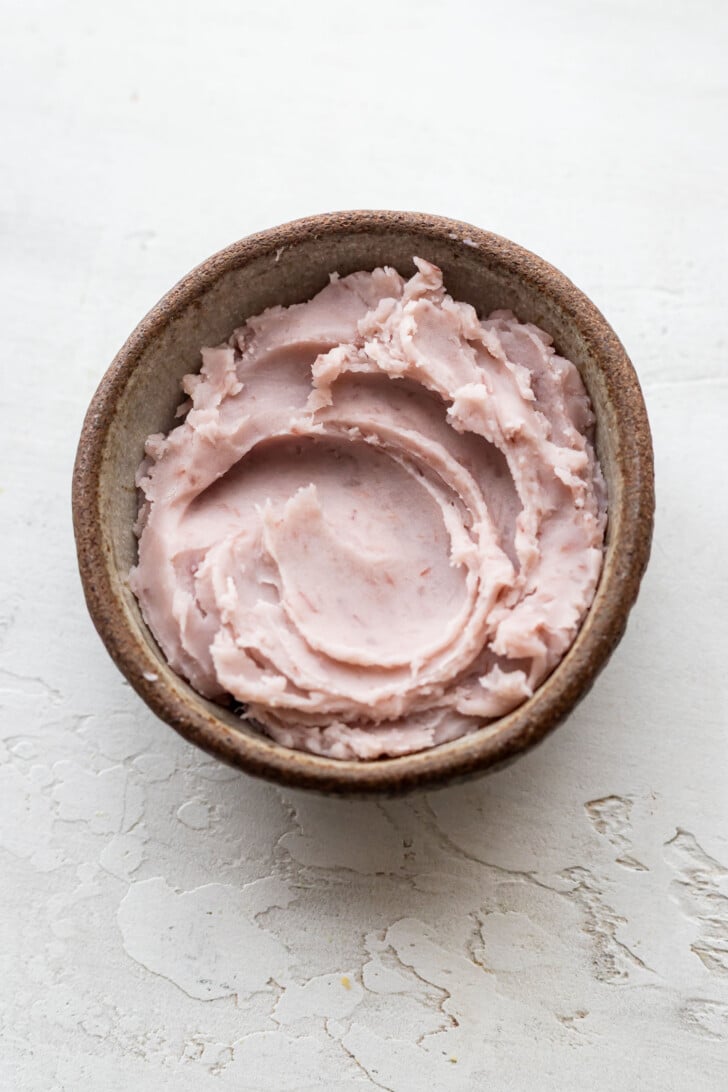 A small brown bowl filled with pink, creamy taro paste spread, set on a textured white surface.