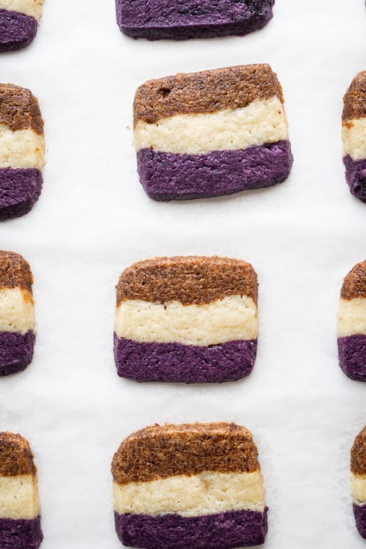 Rectangular cookies with three layers—brown on top, white in the middle, and purple on the bottom—are arranged in rows on white parchment paper.