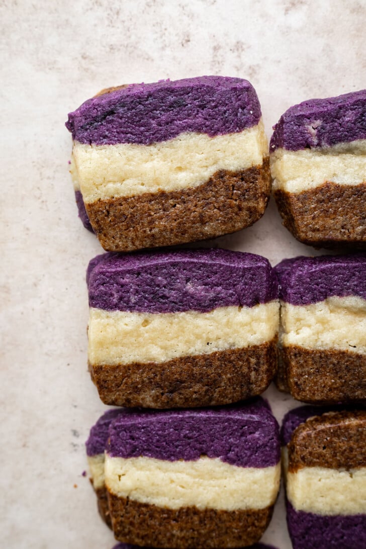 Rectangular ube coffee shortbread cookies with three distinct layers—purple on top, brown on the bottom, and a cream-colored center—are arranged neatly in rows on a flat surface.