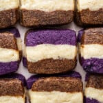Rectangular ube coffee shortbread cookies with three distinct layers—purple on top, brown on the bottom, and a cream-colored center—are arranged neatly in rows on a flat surface.